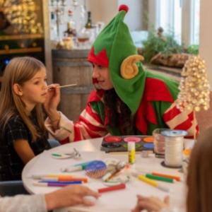 Un elfo dipinge il viso di una bambina durante un'attività creativa natalizia.