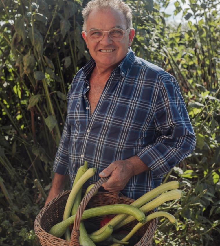 Uomo con camicia a quadri, sorride mentre tiene un cesto di zucchine lunghe.